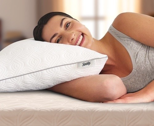 A woman laying in bed and smiling at the camera, as she rests her head on a Sealy Adjustable Pillow